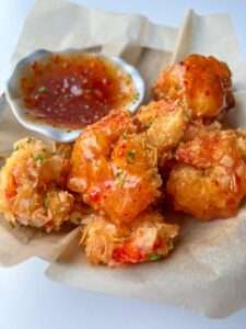 Golden, crispy coconut shrimp pieces—made with our signature Coconut Shrimp Recipe—served on parchment paper with a small dish of sweet chili dipping sauce on the side.