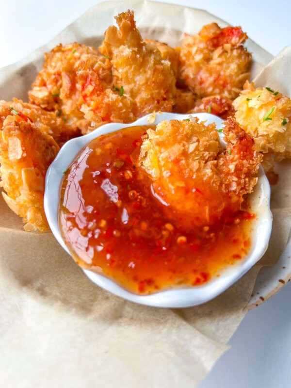 Golden, crispy coconut shrimp—made from our signature Coconut Shrimp Recipe—served in a basket with a small white dish of sweet chili dipping sauce, all nestled on parchment paper.