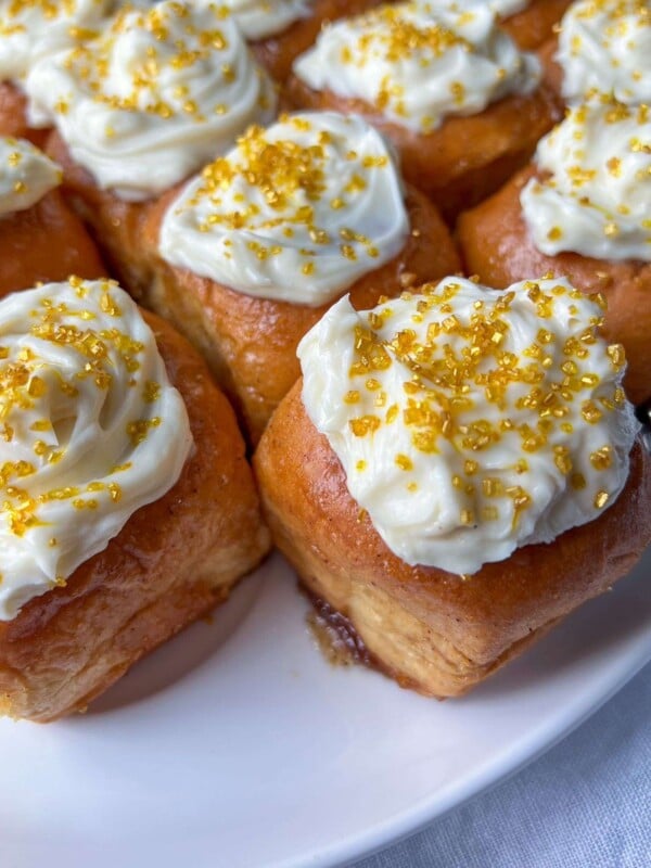 Close-up of golden brown Lazy Cinnamon Buns topped with swirls of white icing and sprinkled with bright yellow-gold sugar crystals, arranged on a white plate.
