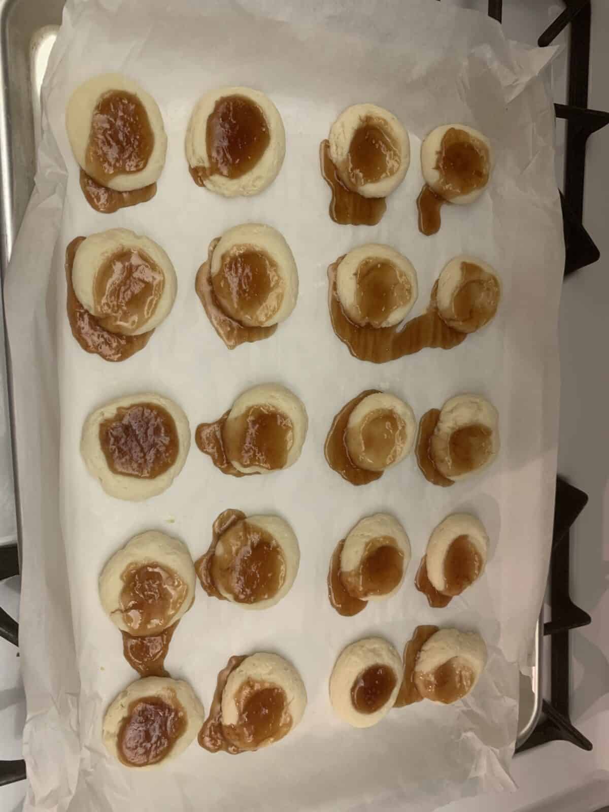 A baking sheet lined with parchment paper holds several unbaked Lemon Cheesecake Thumbprint Cookies filled with caramel or light brown jam, some with filling spilled over the edges, on a stovetop.