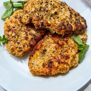 Three pieces of golden-brown, seasoned grilled chicken breast garnished with fresh basil leaves on a white plate.
