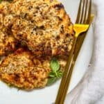 Grilled, seasoned chicken breasts on a white plate garnished with fresh basil, next to a gold fork and knife, set on a white cloth background.