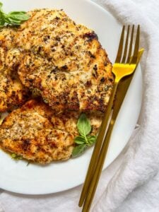 Grilled, seasoned chicken breasts on a white plate garnished with fresh basil, next to a gold fork and knife, set on a white cloth background.