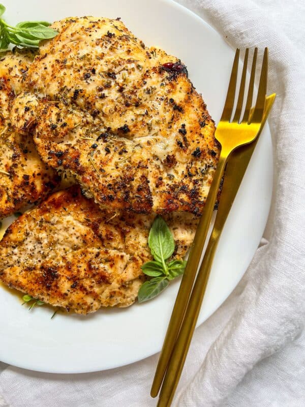 Grilled, seasoned chicken breasts on a white plate garnished with fresh basil, next to a gold fork and knife, set on a white cloth background.