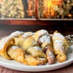 A plate of golden, flaky Easy Nutella Croissants and pastries dusted with powdered sugar sits on a table, with a blurred warm-toned background of festive lights.