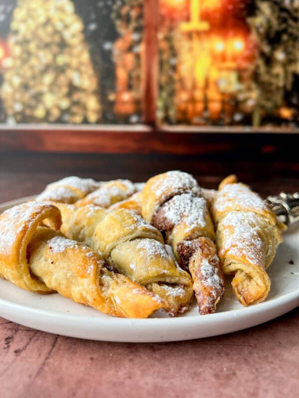 A plate of golden, flaky Easy Nutella Croissants and pastries dusted with powdered sugar sits on a table, with a blurred warm-toned background of festive lights.