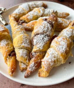A plate of golden-brown Easy Nutella Croissants dusted with powdered sugar, arranged closely together. The pastries look flaky and slightly crisp with a hint of rich chocolate filling visible.