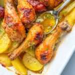 Roasted Chicken with Ribs drumsticks, golden potato slices, and fresh herbs fill a white baking dish, while a spoon serves the delicious food.