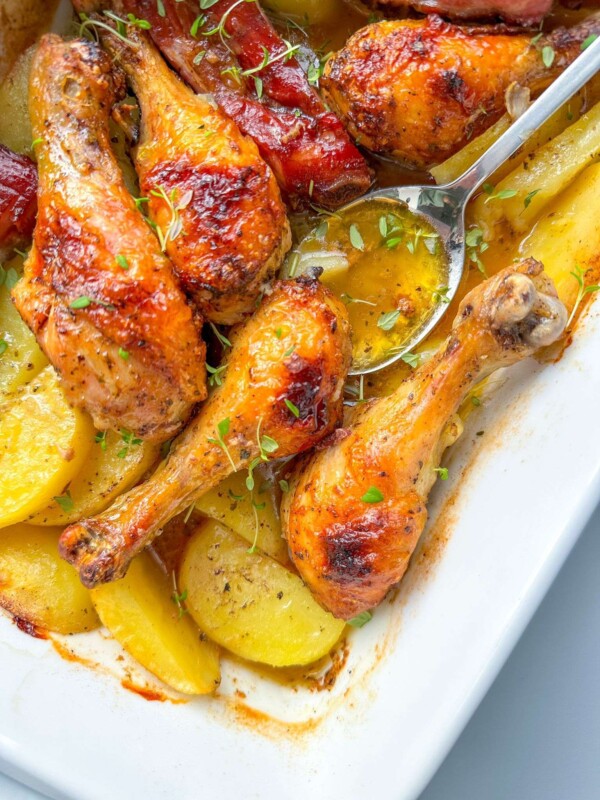 Roasted Chicken with Ribs drumsticks, golden potato slices, and fresh herbs fill a white baking dish, while a spoon serves the delicious food.