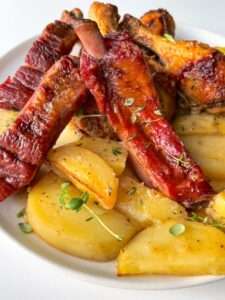 A plate filled with Roasted Chicken with Ribs, glazed pork ribs, and golden roasted potato wedges, all garnished with fresh herbs.