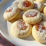 A plate of thumbprint cookies, reminiscent of Shortbread Cookie Bars, topped with powdered sugar and filled with orange jam, arranged in sunlight on a white dish.