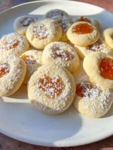 A plate of thumbprint cookies filled with orange jam and dusted with powdered sugar, these treats offer the same buttery delight as Shortbread Cookie Bars, beautifully arranged in sunlight on a white plate.