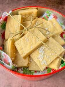 A tin filled with rectangular Shortbread Cookie Bars, some topped with white sprinkles, tied together with a gold string, and resting on festive holiday-themed paper.