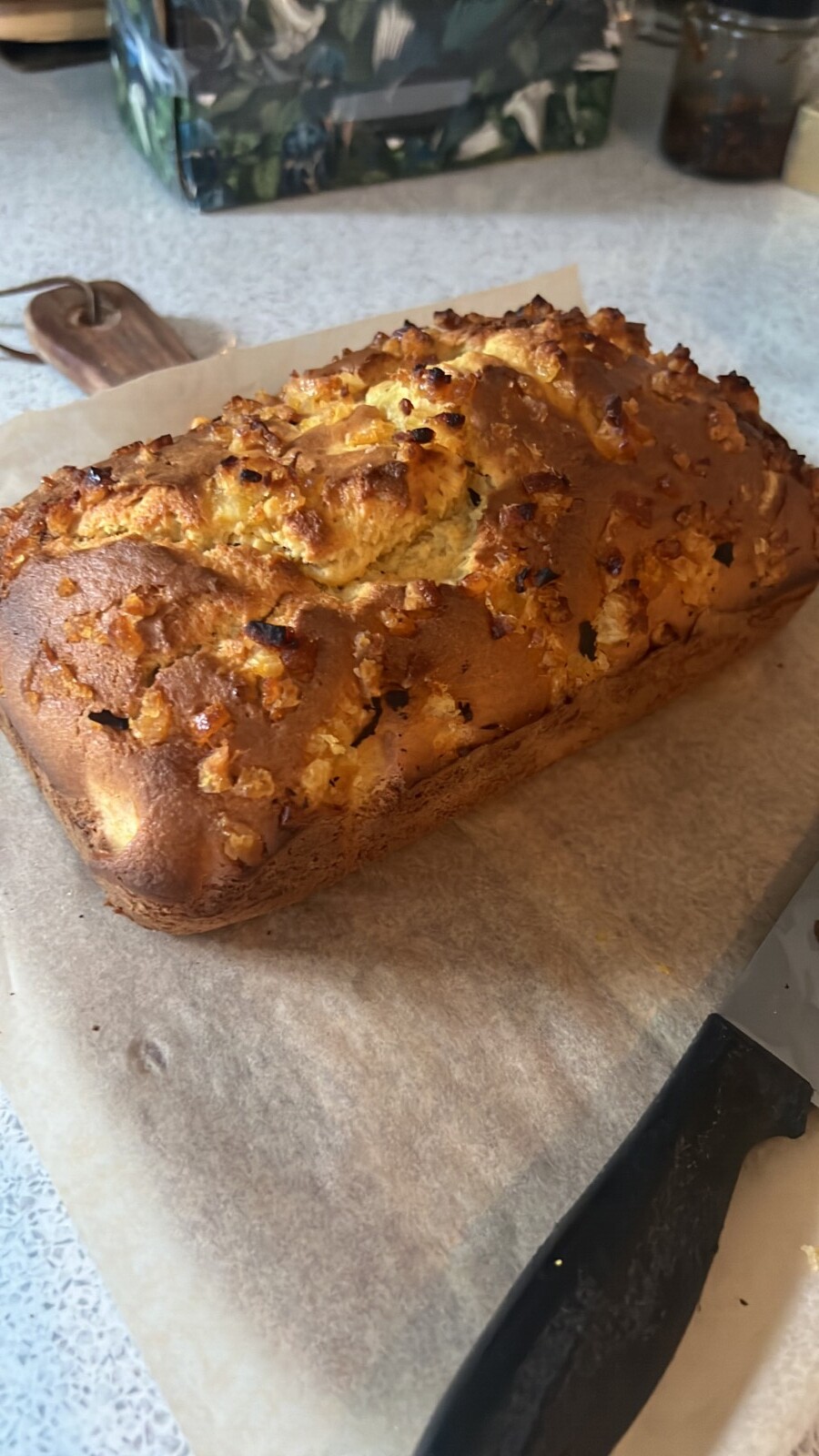 A freshly baked loaf of Whole Orange Blender Cake with a golden-brown crust and crunchy topping sits on parchment paper next to a knife on a kitchen counter.