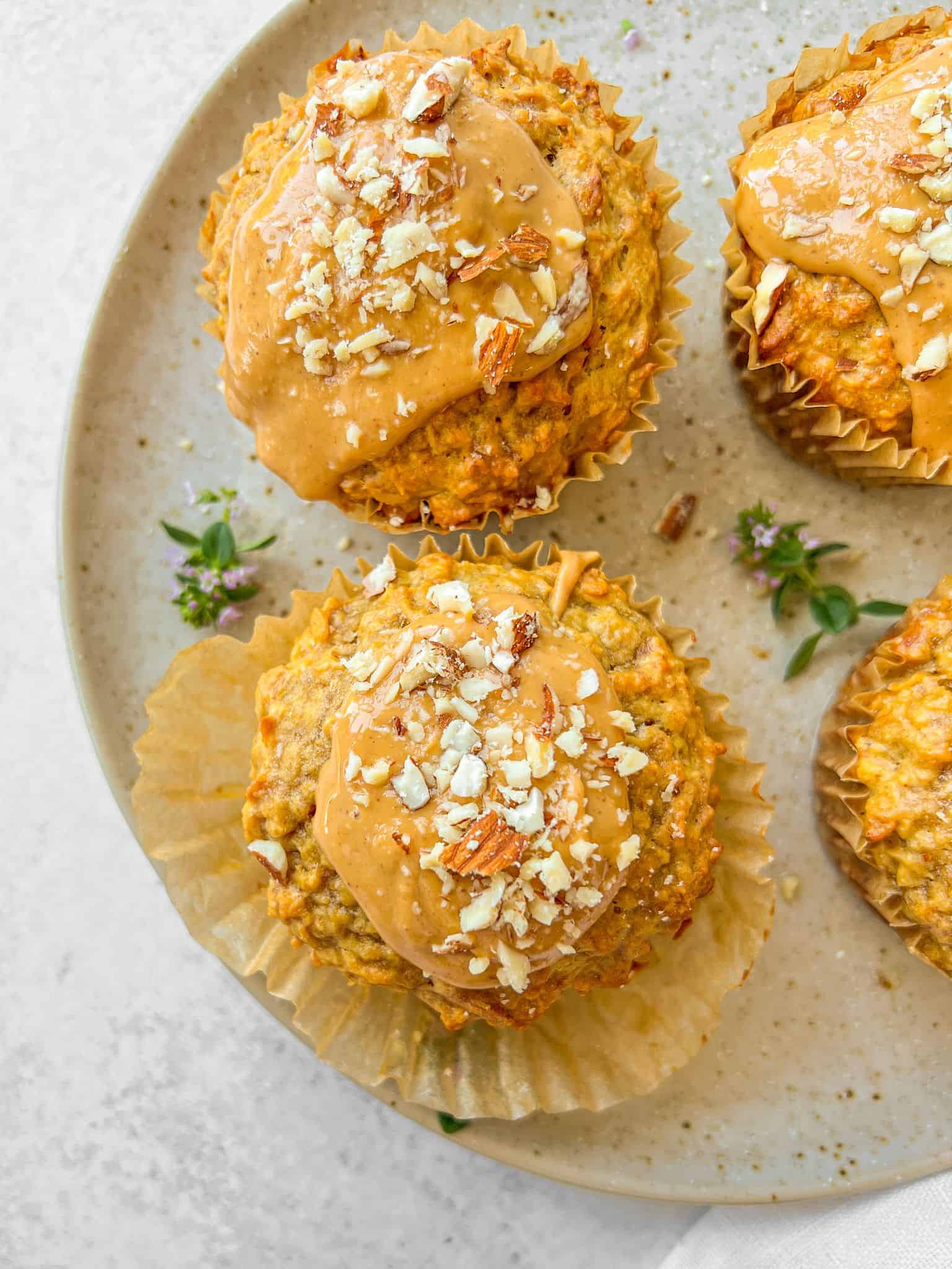 Three muffins topped with a spread and chopped nuts sit on a speckled plate. The muffins are in crinkled paper liners, and small purple flowers are scattered on the plate. The background is a light gray surface.