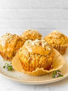 Four muffins on a plate, each topped with a glossy spread and sprinkled with chopped nuts. The plate is garnished with small purple flowers. The background is a white brick wall, adding a rustic touch.