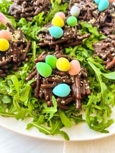 Chocolate nests topped with colorful candy eggs sit on a bed of fresh arugula. The nests are made of chocolate-coated sticks and sprinkled with sesame seeds. The eggs are pastel shades of blue, green, yellow, and pink.