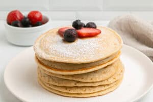A stack of pancakes dusted with powdered sugar, topped with sliced strawberries and blueberries, on a white plate. A small bowl of mixed berries is in the background.