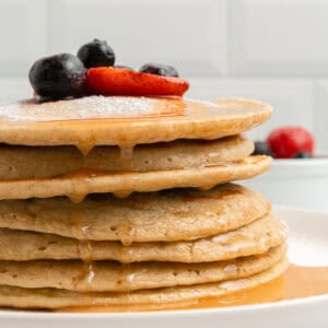 A stack of pancakes drizzled with syrup, topped with powdered sugar, blueberries, and sliced strawberries on a white plate. A bowl of berries is blurred in the background.