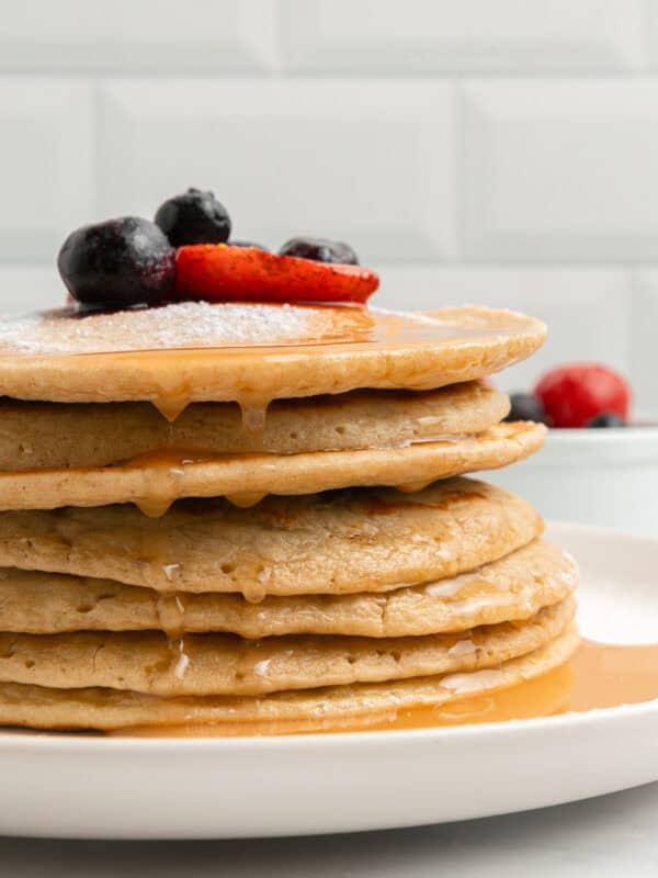 A stack of pancakes drizzled with syrup, topped with powdered sugar, blueberries, and sliced strawberries on a white plate. A bowl of berries is blurred in the background.