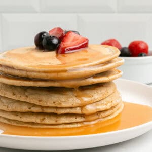 A stack of pancakes topped with sliced strawberries and blueberries, drizzled with syrup, on a white plate. In the background, a small bowl of fresh berries sits on a white surface.