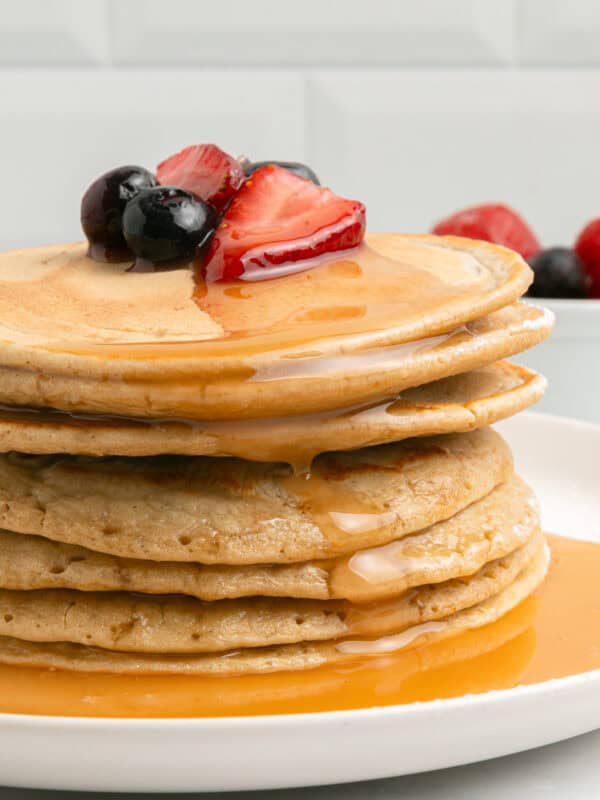 A stack of pancakes topped with sliced strawberries and blueberries, drizzled with syrup, on a white plate. In the background, a small bowl of fresh berries sits on a white surface.