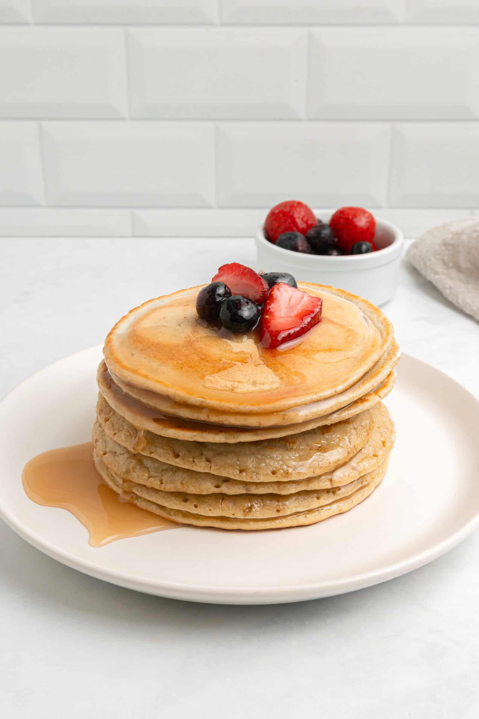A stack of pancakes topped with strawberries and blueberries, drizzled with syrup, sits on a white plate. Extra syrup pools on the plate, and a small bowl of mixed berries is in the background.