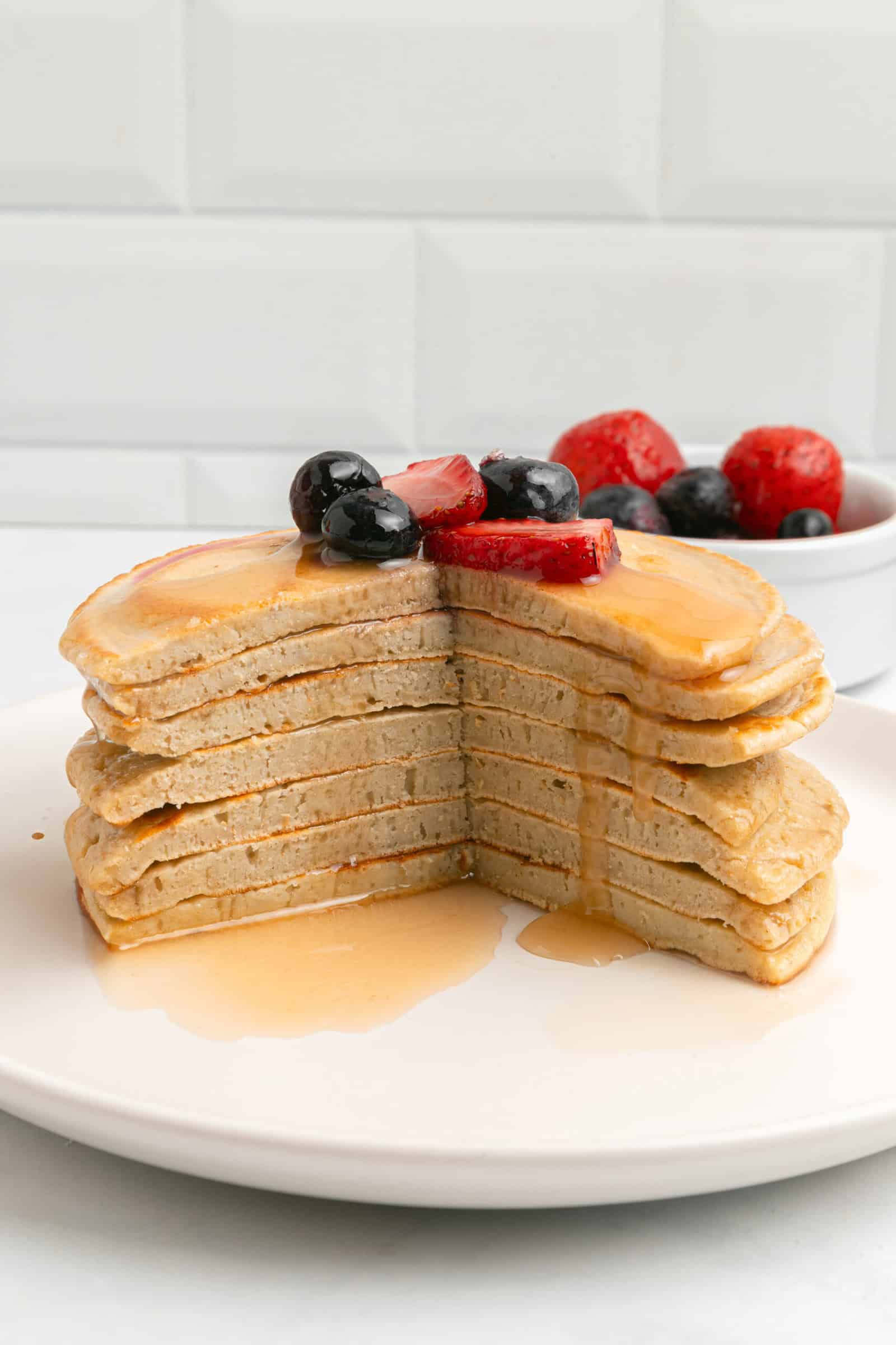 A stack of pancakes with a large wedge cut out, topped with syrup and fresh blueberries and strawberries. A small bowl of mixed berries is in the background.