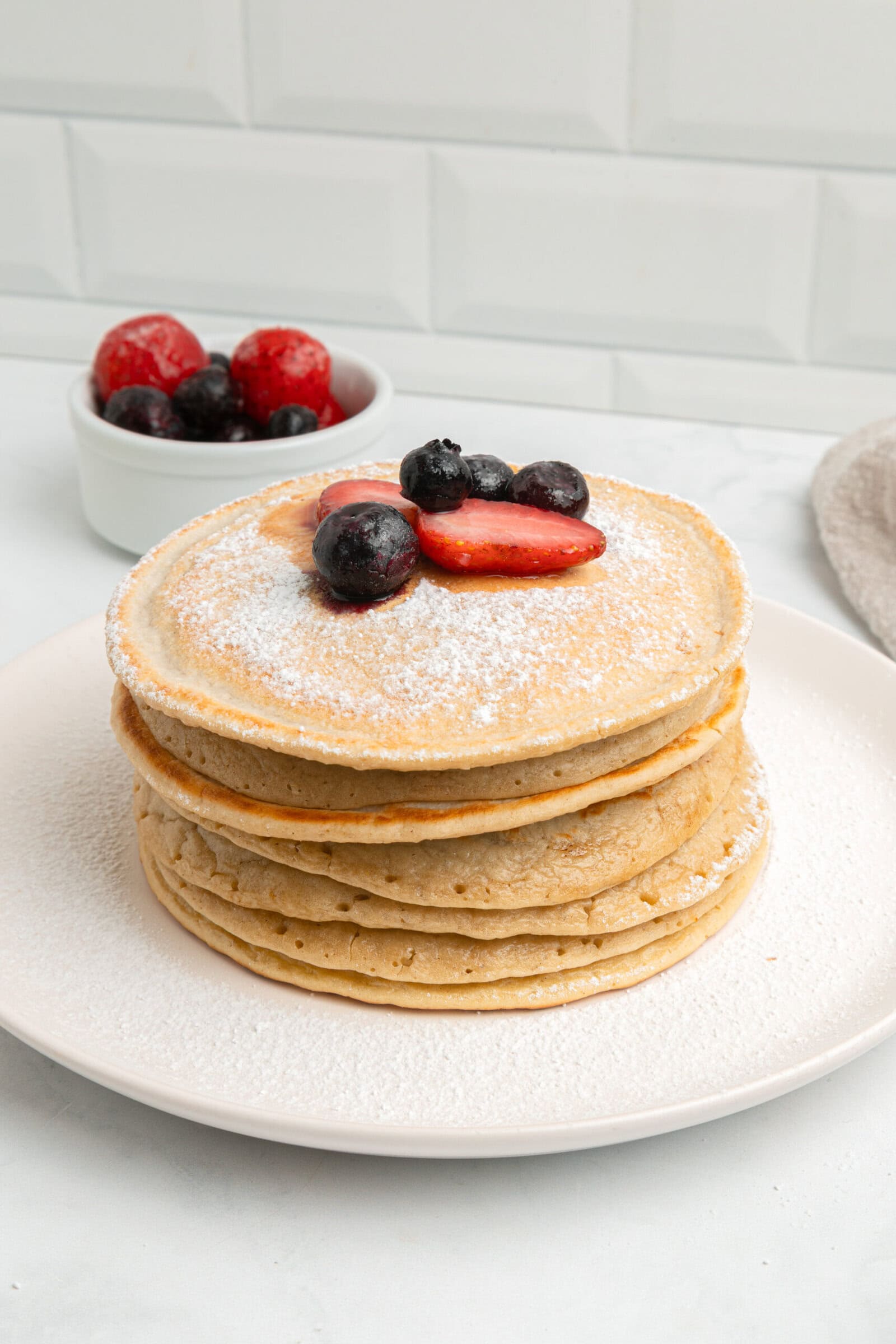 A stack of pancakes topped with powdered sugar, strawberries, and blueberries on a white plate, with a small bowl of mixed berries in the background.