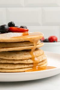 A stack of pancakes topped with sliced strawberries and blueberries, with syrup dripping down the sides, served on a white plate. A bowl of assorted berries is blurred in the background.