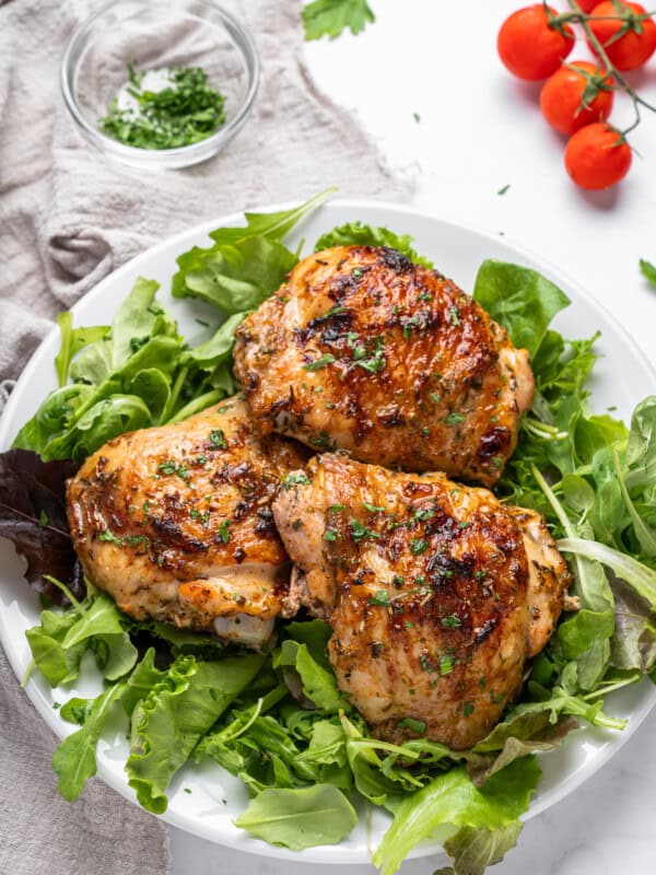 Grilled chicken thighs garnished with herbs are served on a bed of mixed greens on a white plate. A small bowl of chopped herbs and a bunch of fresh cherry tomatoes are in the background.