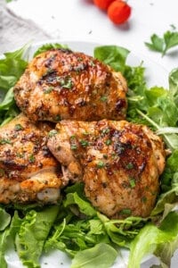 Grilled chicken thighs with a golden, crispy skin are garnished with herbs and served on a bed of fresh leafy greens. A few ripe red tomatoes can be seen in the background.