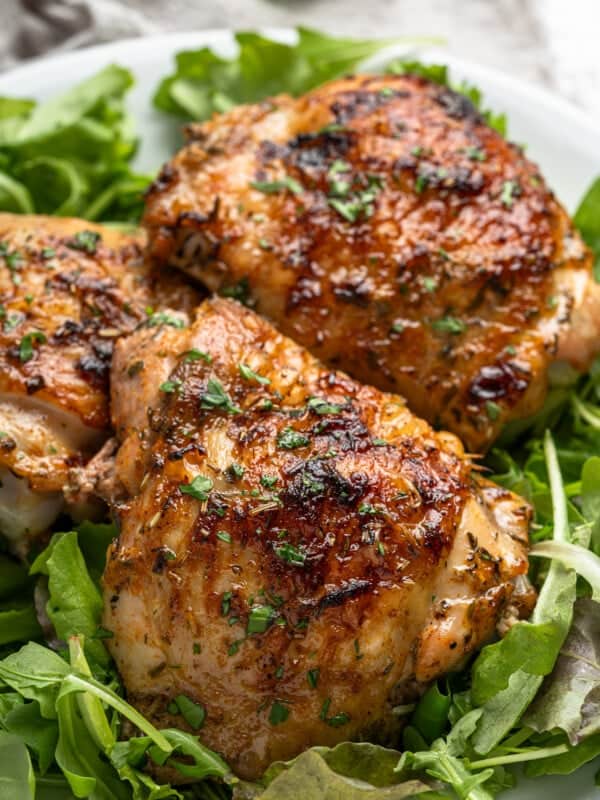 Grilled chicken thighs garnished with herbs are served on a bed of fresh arugula. The chicken is golden-brown and appears juicy and flavorful, sitting on a white plate.