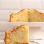 A close-up of a slice of apple cake on a plate, with the rest of the cake on a stand in the background. The cake appears moist and dense, with visible apple pieces. White tiled wall in the background.