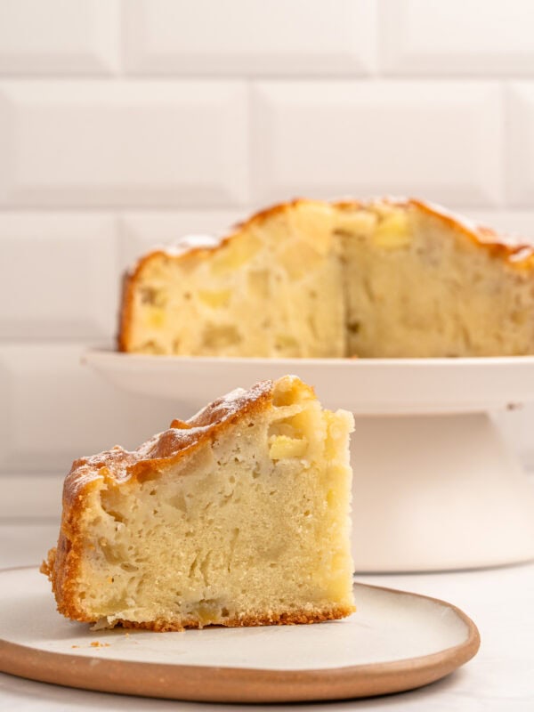 A close-up of a slice of apple cake on a plate, with the rest of the cake on a stand in the background. The cake appears moist and dense, with visible apple pieces. White tiled wall in the background.