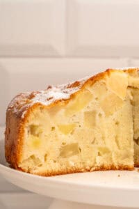 A close-up of a sliced apple cake on a white plate, showing a moist, dense crumb with visible apple chunks and a light dusting of powdered sugar on top.