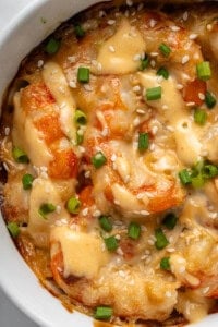 Close-up of a dish featuring plump, cooked shrimp covered in a creamy, golden sauce. The shrimp are garnished with chopped green onions and sesame seeds, all served in a white bowl.