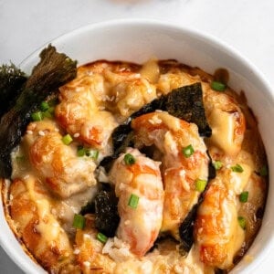 A bowl of shrimp gratin topped with melted cheese, seaweed, and green onions. The dish is garnished with a sprinkle of sesame seeds. A small bowl of soy sauce is in the background on a white marble surface.