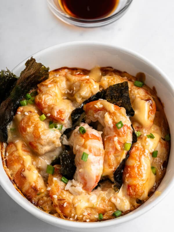 A bowl of shrimp gratin topped with melted cheese, seaweed, and green onions. The dish is garnished with a sprinkle of sesame seeds. A small bowl of soy sauce is in the background on a white marble surface.