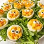 Deviled eggs decorated to resemble chicks with black olive eyes and carrot beaks, arranged on a bed of arugula on a white plate.