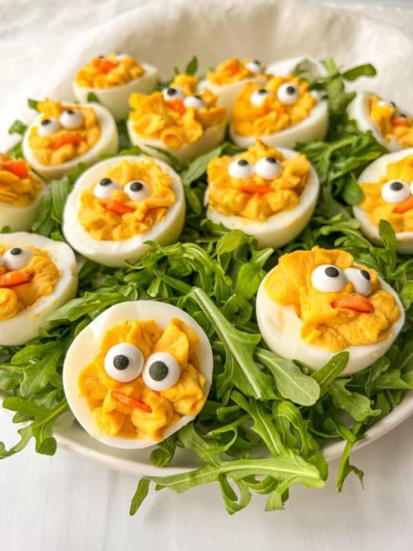 Deviled eggs decorated to resemble chicks with black olive eyes and carrot beaks, arranged on a bed of arugula on a white plate.