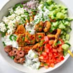 An Easy Greek Bowl filled with grilled chicken, diced cucumber, tomato, red onion, olives, feta cheese, and a drizzle of creamy white dressing, all garnished with chopped parsley.