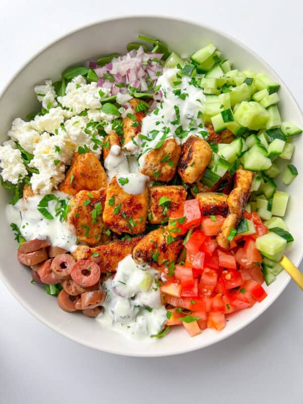 An Easy Greek Bowl filled with grilled chicken, diced cucumber, tomato, red onion, olives, feta cheese, and a drizzle of creamy white dressing, all garnished with chopped parsley.