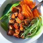 A vibrant bowl inspired by a Honey Garlic Salmon Bowl, featuring glazed tofu cubes, shredded carrots, spiralized cucumber, fresh spinach, rice, and a sprinkle of sesame seeds, served with chopsticks.