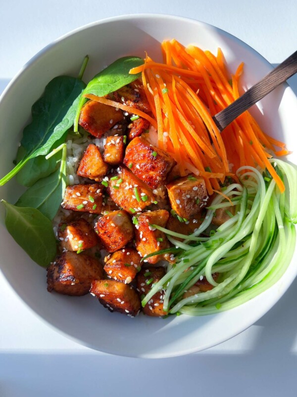 A vibrant bowl inspired by a Honey Garlic Salmon Bowl, featuring glazed tofu cubes, shredded carrots, spiralized cucumber, fresh spinach, rice, and a sprinkle of sesame seeds, served with chopsticks.