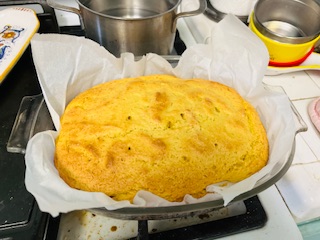 A freshly baked golden-brown cornbread sits in a parchment-lined pan on a stove, with pots and kitchen utensils nearby—evoking the comforting aroma of a Whole Orange Blender Cake just out of the oven.