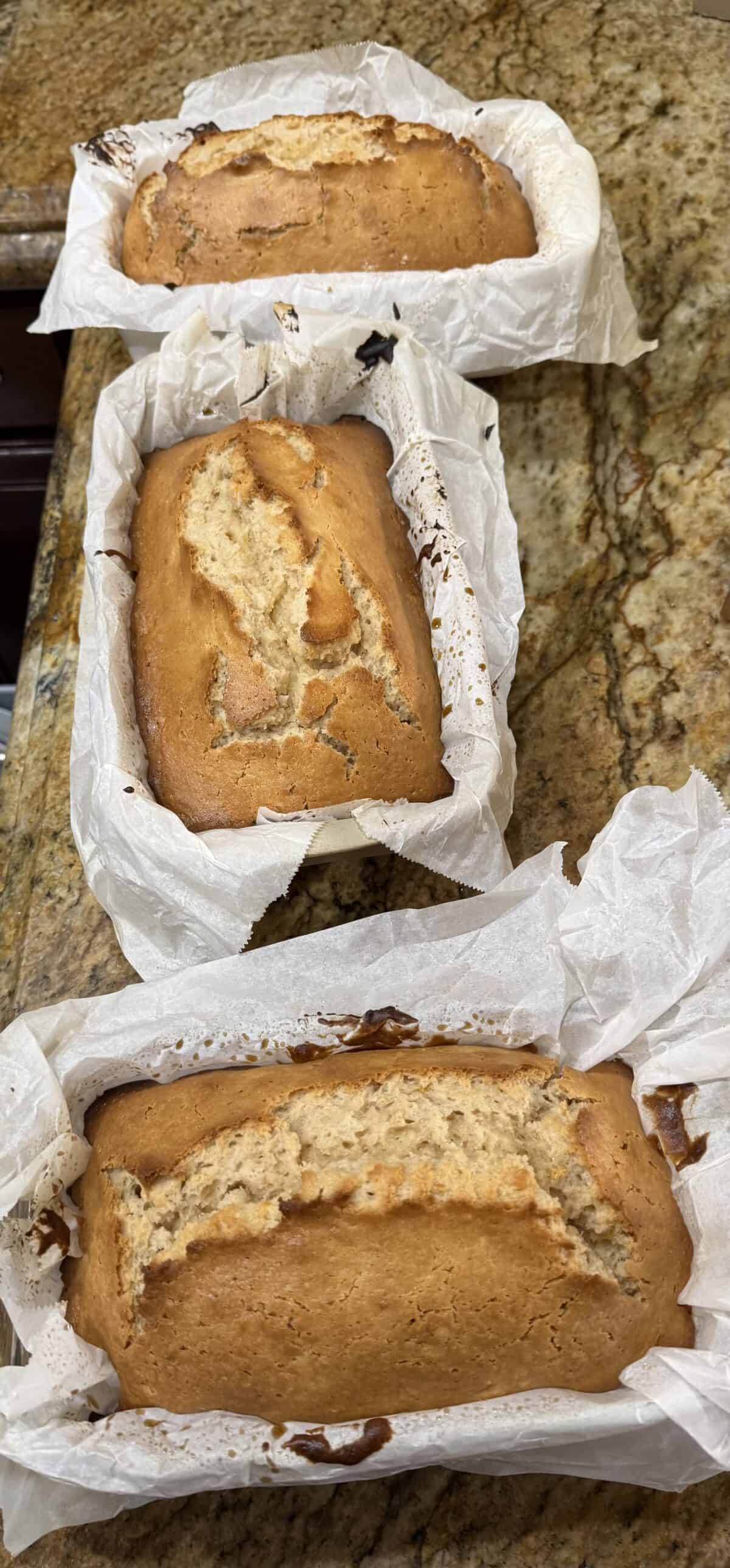 Three freshly baked loaves of Whole Orange Blender Cake sit in parchment-lined pans on a marbled kitchen countertop. The cakes have golden brown crusts and visible cracks on top.