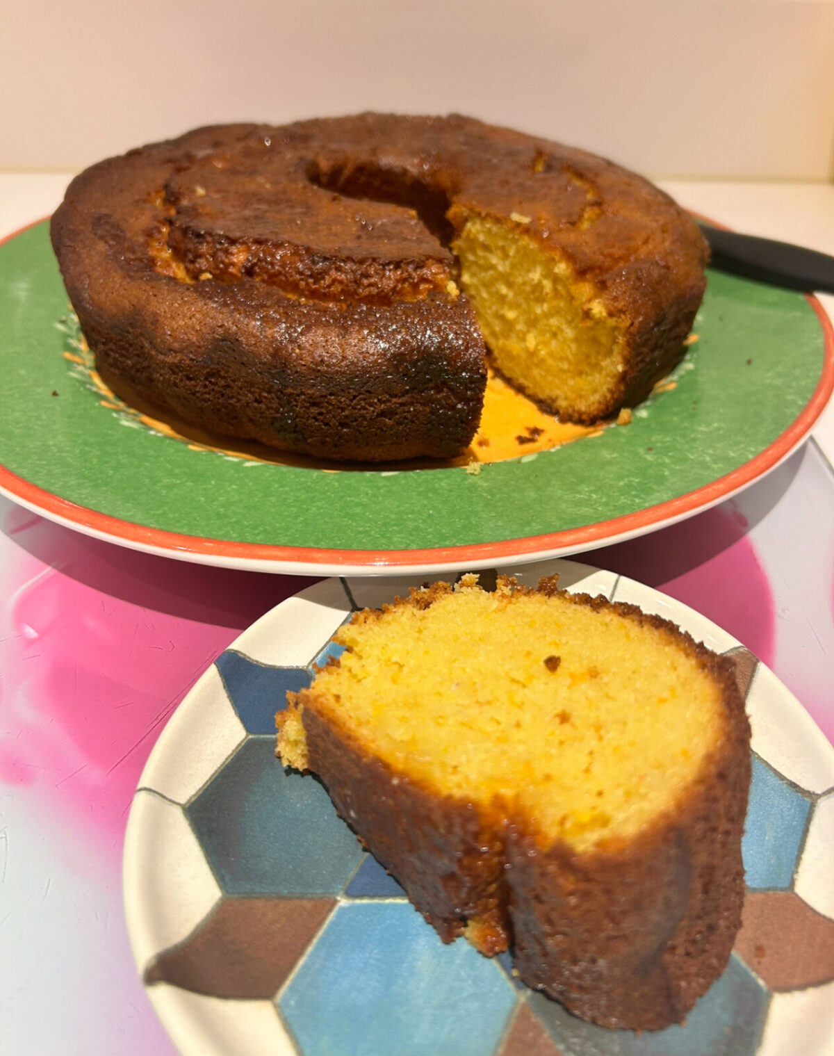 A round, golden-brown Whole Orange Blender Cake with a slice cut out sits on a green plate in the background. In the foreground, the slice rests on a small, patterned plate, revealing the cake’s moist yellow interior.