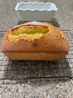 A golden-brown loaf of Whole Orange Blender Cake sits cooling on a wire rack atop a speckled countertop, with a white ceramic loaf pan in the background.
