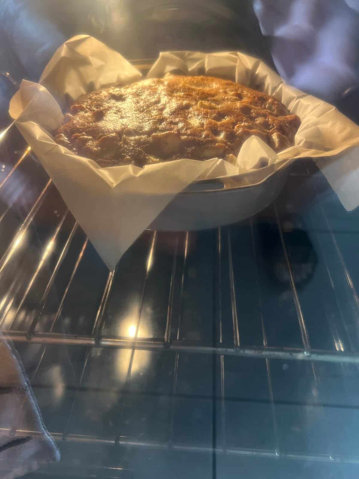 A round cake in a parchment-lined pan is baking in an oven, with the cake rising and browning on top. The oven rack and some reflections on the oven door are visible.